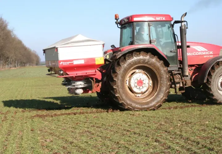 Czerwony traktor McCormick z rozsiewaczem na polu. Rozsiewacz rozprowadza saletrę wapniową, kluczową dla zdrowia roślin.