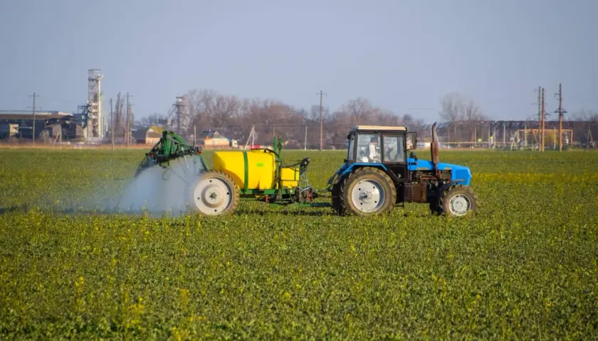 Niebieski traktor z żółtym zbiornikiem wykonuje oprysk na polu zielonych roślin. W tle widać przemysłowe budynki.