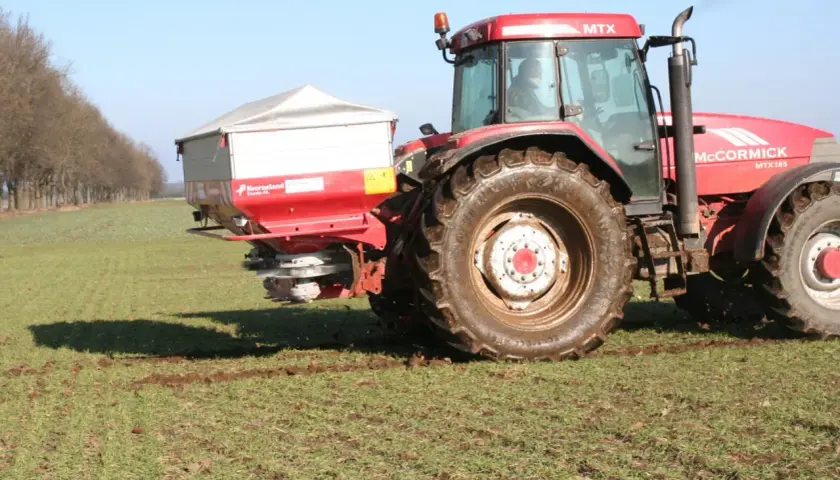Czerwony traktor McCormick z rozsiewaczem na polu. Rozsiewacz rozprowadza saletrę wapniową, kluczową dla zdrowia roślin.