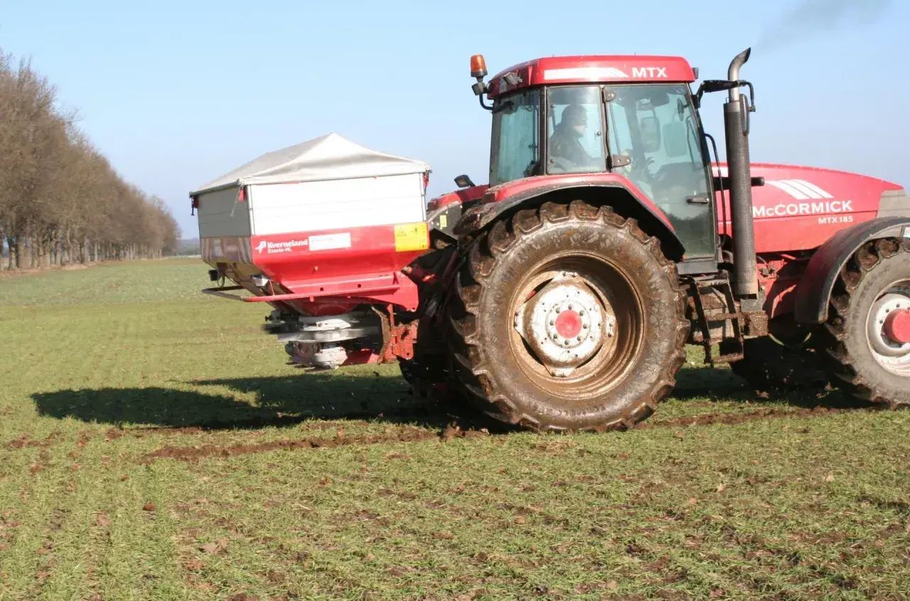 Czerwony traktor McCormick z rozsiewaczem na polu. Rozsiewacz rozprowadza saletrę wapniową, kluczową dla zdrowia roślin.